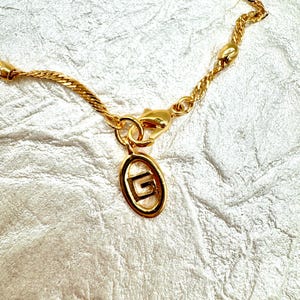 May include: A gold-toned necklace with a delicate chain and a pendant. The pendant is an oval shape with a letter "G" inside. The necklace has a lobster clasp closure. The background is a textured, off-white surface.