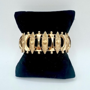May include: A gold-tone metal bracelet featuring a repeating pattern of alternating vertical marquise-shaped links with a brushed texture and smooth, curved concave links. Each marquise link is accented with a small central sphere and ball ends.