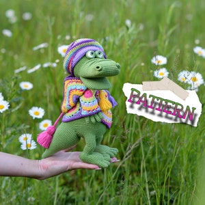 Può includere: Un alligatore verde all'uncinetto con un maglione a righe colorato, un cappello e una sciarpa con un nappa rosa. L'alligatore è tenuto in mano in un campo di margherite. La parola "PATTERN" è nell'immagine.