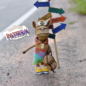 May include: A brown crocheted camel toy with a rainbow striped shirt and a small backpack. The camel is standing on a road with a signpost pointing to different cities around the world, including Moscow, Bangkok, Madrid, Rome, Paris, Manila, Lima, and Tokyo. The signpost is made of wood and has a brown paper sign that says "PATTERN".