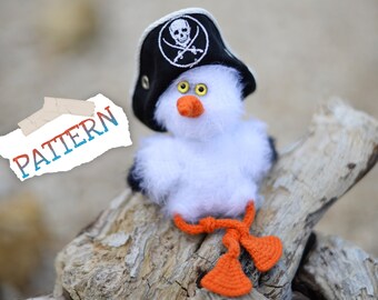 May include: A white crocheted bird wearing a black pirate hat with a skull and crossbones design. The bird is sitting on a piece of driftwood.