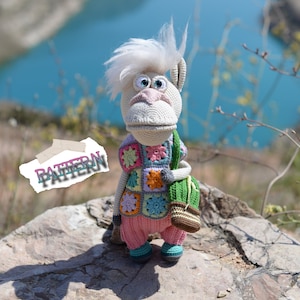 May include: A crocheted stuffed toy with white fur, pink trousers, and a colourful patchwork waistcoat. The toy has a long snout, large eyes, and a green bag.