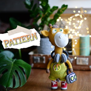 May include: Crocheted giraffe toy wearing headphones and holding a small radio. The giraffe is yellow and brown with a white face and has a peace sign on its overalls. The word "PATTERN" is written on a piece of paper in the background.
