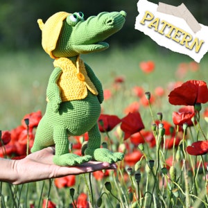 May include: A green crocheted crocodile wearing a yellow hat and vest. The crocodile is standing on a hand in a field of red poppies.