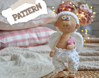 May include: A crocheted angel doll with a halo, white wings, and a pink heart in its hands. The doll is sitting on a white blanket with a pink and white checkered pillow behind it.