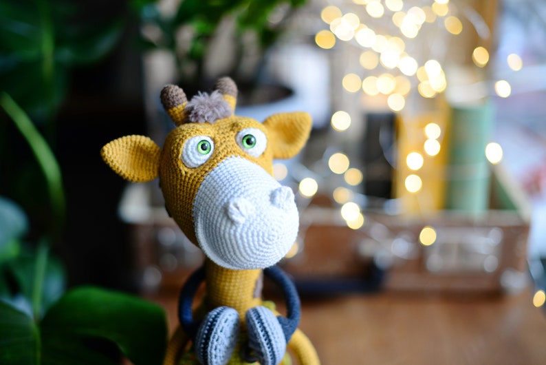 May include: A crocheted giraffe toy with large green eyes, a white crocheted nose, and yellow and brown fur. The giraffe is wearing a blue and gray crocheted scarf.