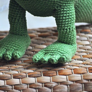 May include: Close-up of a green crocheted dinosaur toy with a woven texture. The dinosaur is standing on a brown woven mat.