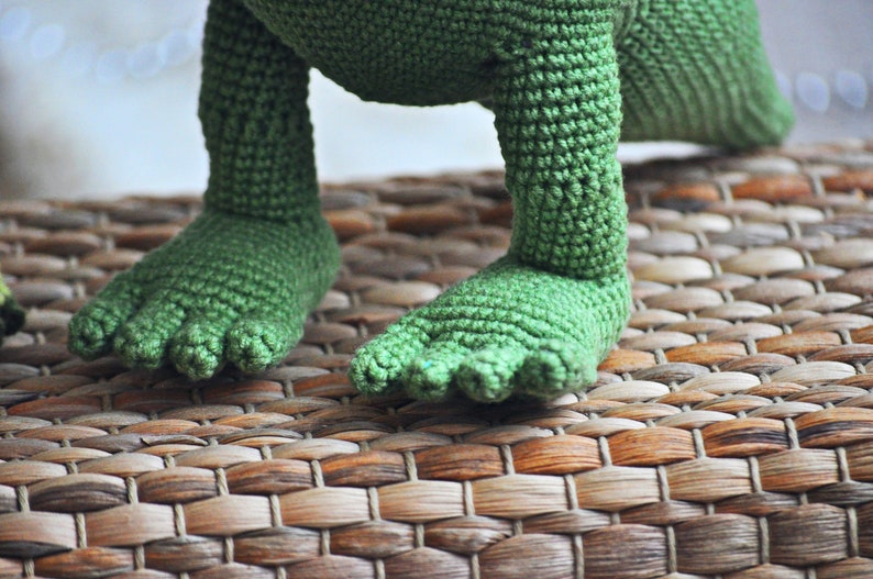May include: Close-up of a green crocheted dinosaur toy with a woven texture. The dinosaur is standing on a brown woven mat.