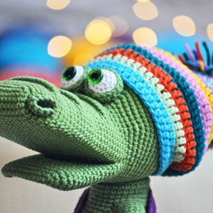 May include: Green crocheted alligator wearing a colorful striped hat with a purple pom-pom.
