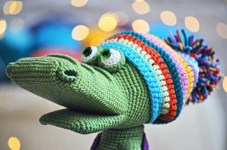 May include: Green crocheted alligator wearing a colorful striped hat with a purple pom-pom.