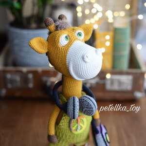 May include: A crocheted giraffe toy wearing headphones and a green overall with a peace sign. The giraffe is holding a purple and green crocheted bag. The toy is standing on a wooden surface.
