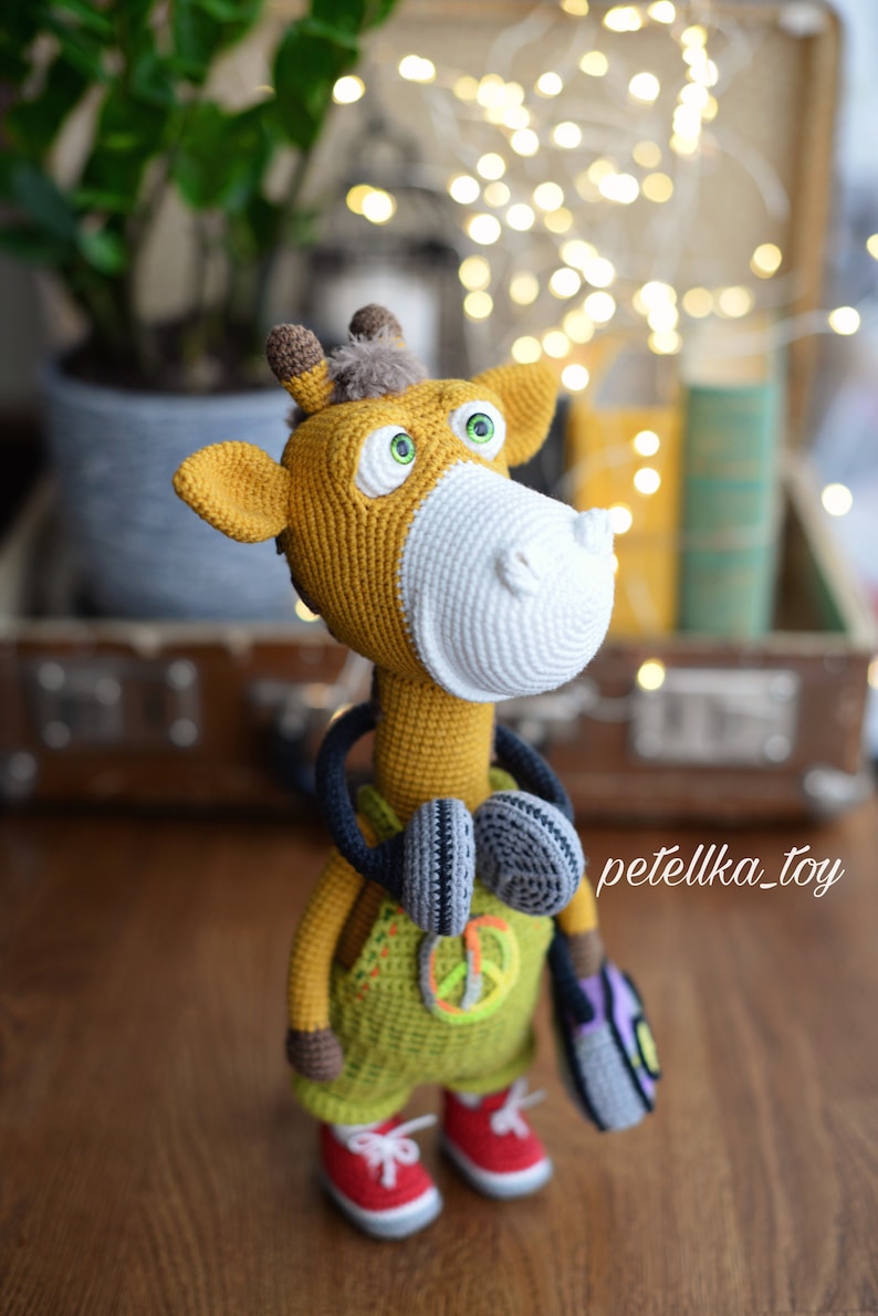 May include: A crocheted giraffe toy wearing headphones and a green overall with a peace sign. The giraffe is holding a purple and green crocheted bag. The toy is standing on a wooden surface.