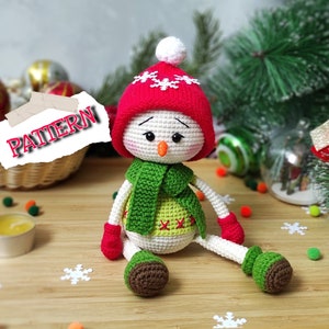Op de afbeelding: Een gehaakt sneeuwpop speelgoed dat een rode muts met witte sneeuwvlokken, een groene sjaal en groene wanten draagt. De sneeuwpop zit op een houten oppervlak met een kerstboom op de achtergrond.