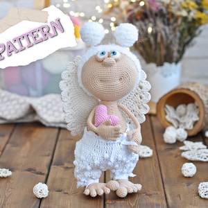 Könnte beinhalten: Eine gehäkelte Amigurumi-Engelpuppe mit weißen Flügeln, einem weißen Kleid und einem rosa Herzen. Die Puppe trägt einen weißen Häkelhut mit zwei weißen Bommeln oben. Die Puppe hält ein rosa Herz in ihren Händen.