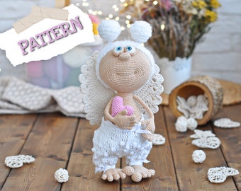 May include: A crocheted amigurumi angel doll with white wings, a white dress, and a pink heart. The doll has a white crocheted hat with two white pom-poms on top. The doll is holding a pink heart in its hands.