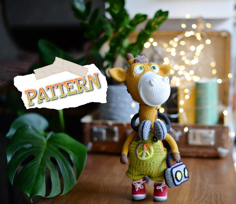 May include: Crocheted giraffe toy wearing headphones and holding a small radio. The giraffe is yellow and brown with a white face and has a peace sign on its overalls. The word "PATTERN" is written on a piece of paper in the background.