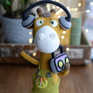 May include: A crocheted giraffe toy wearing headphones and holding a small radio. The giraffe is wearing green overalls with a peace sign on the front. The toy is standing on a wooden surface.