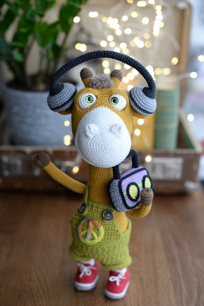 May include: A crocheted giraffe toy wearing headphones and holding a small radio. The giraffe is wearing green overalls with a peace sign on the front. The toy is standing on a wooden surface.