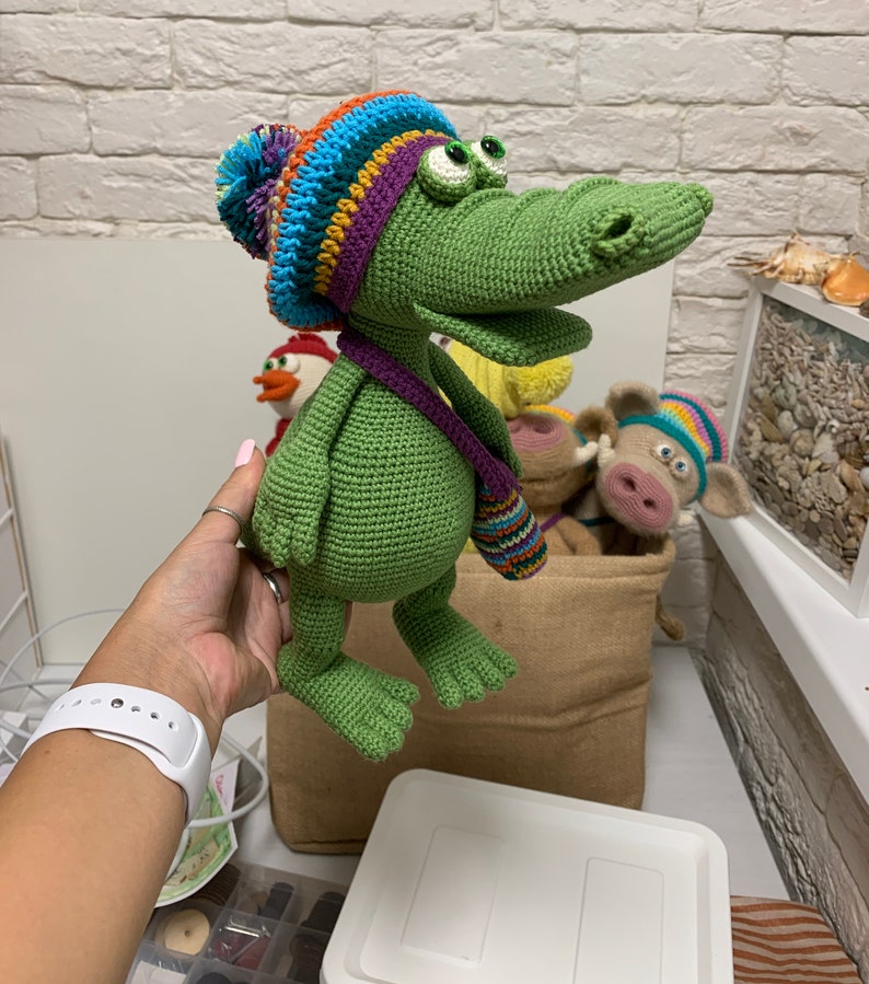 May include: A green crocheted alligator wearing a colorful crocheted hat with a pom-pom. The alligator is holding a small crocheted bag with colorful stripes.