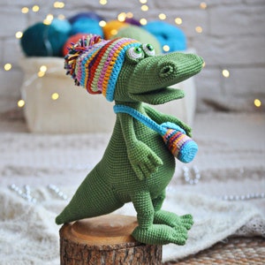 May include: A green crocheted dinosaur wearing a colorful striped hat and carrying a small crocheted bag. The dinosaur is standing on a wooden stump.