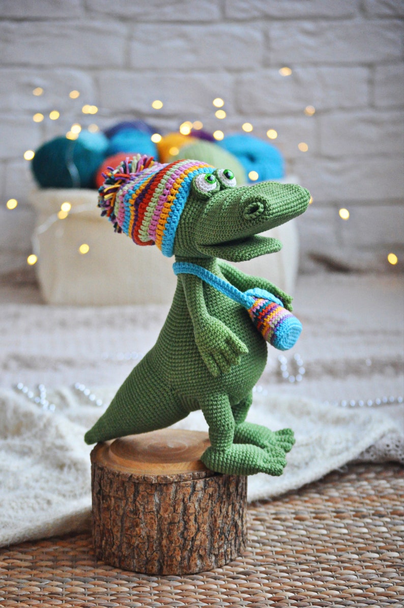 May include: A green crocheted dinosaur wearing a colorful striped hat and carrying a small crocheted bag. The dinosaur is standing on a wooden stump.