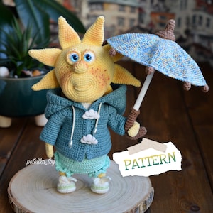 May include: A crocheted sun toy wearing a blue hooded jacket with white cloud buttons, holding a blue and white crocheted umbrella. The sun has a yellow face with orange freckles and blue eyes. The toy is standing on a wooden surface. The text "PATTERN" is written on a piece of paper in the image.