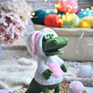 May include: A green crocheted alligator wearing a white crocheted sweater with pink bunny ears and a pink heart on the front. The alligator is standing on a woven mat with white pom-poms.