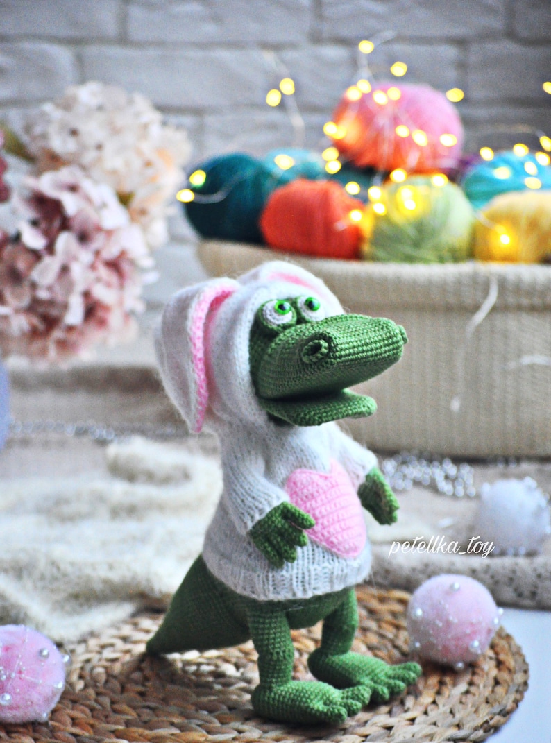 May include: A green crocheted alligator wearing a white crocheted sweater with pink bunny ears and a pink heart on the front. The alligator is standing on a woven mat with white pom-poms.