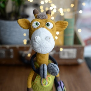 May include: A crocheted giraffe toy wearing a green jumpsuit with a peace sign, headphones, and a purple bag. The giraffe has large green eyes and a brown nose.