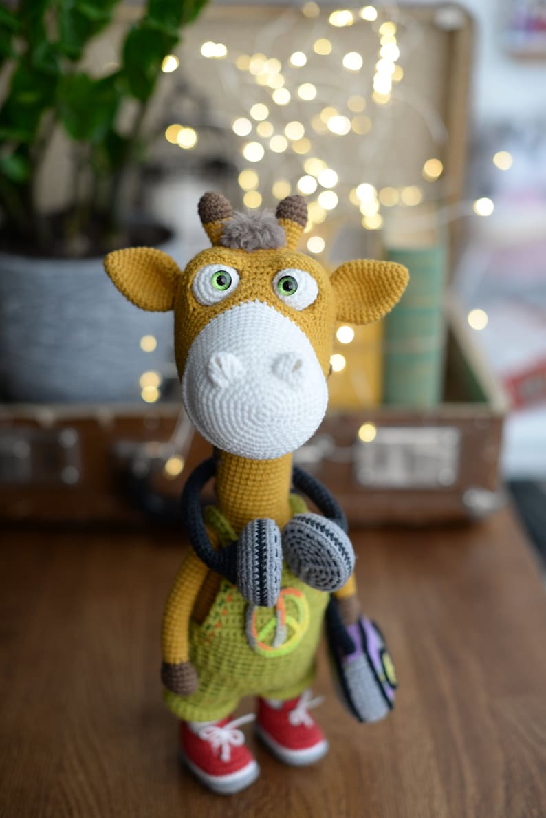May include: A crocheted giraffe toy wearing a green jumpsuit with a peace sign, headphones, and a purple bag. The giraffe has large green eyes and a brown nose.