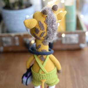 May include: A crocheted giraffe toy wearing overalls and carrying a small purple bag. The giraffe has a brown and yellow patterned body and a fluffy brown tail.