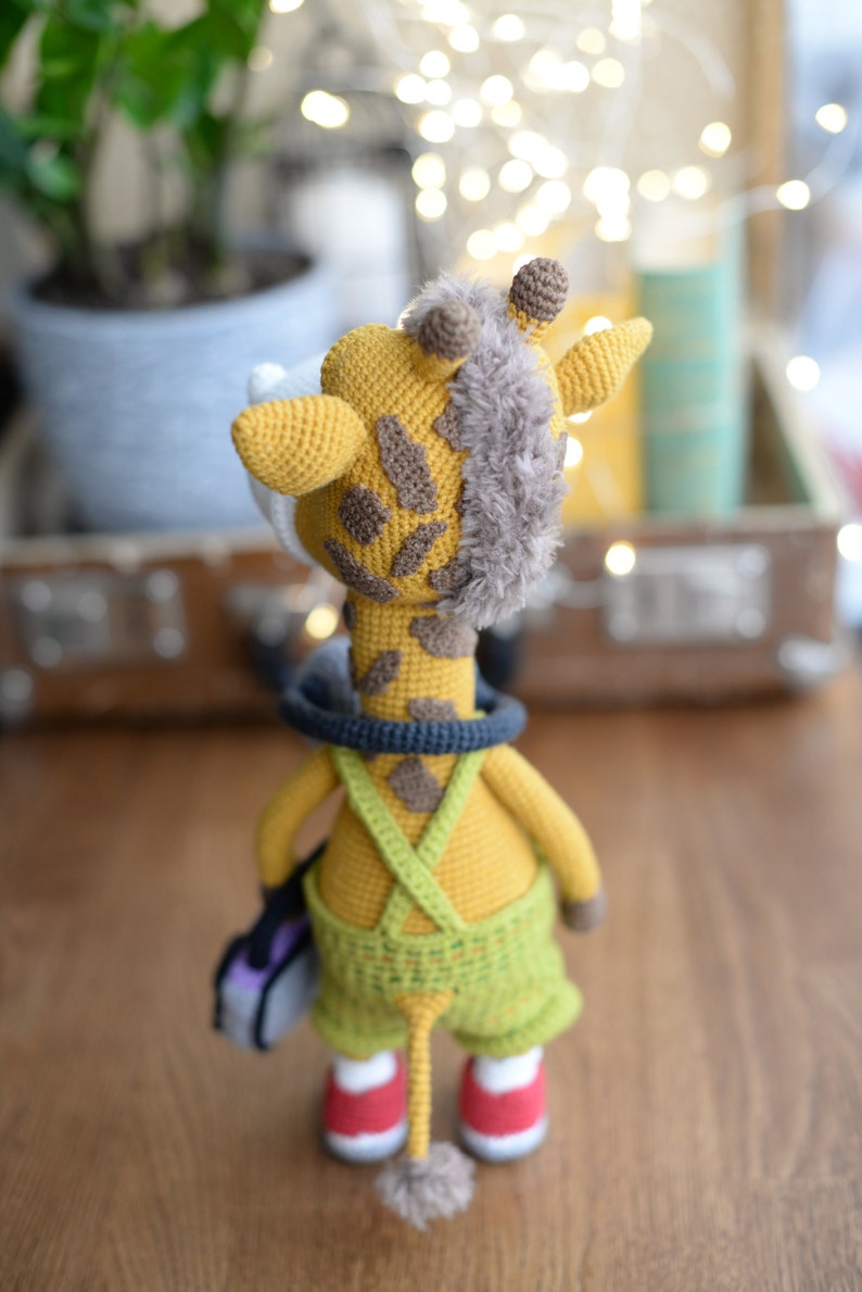 May include: A crocheted giraffe toy wearing overalls and carrying a small purple bag. The giraffe has a brown and yellow patterned body and a fluffy brown tail.