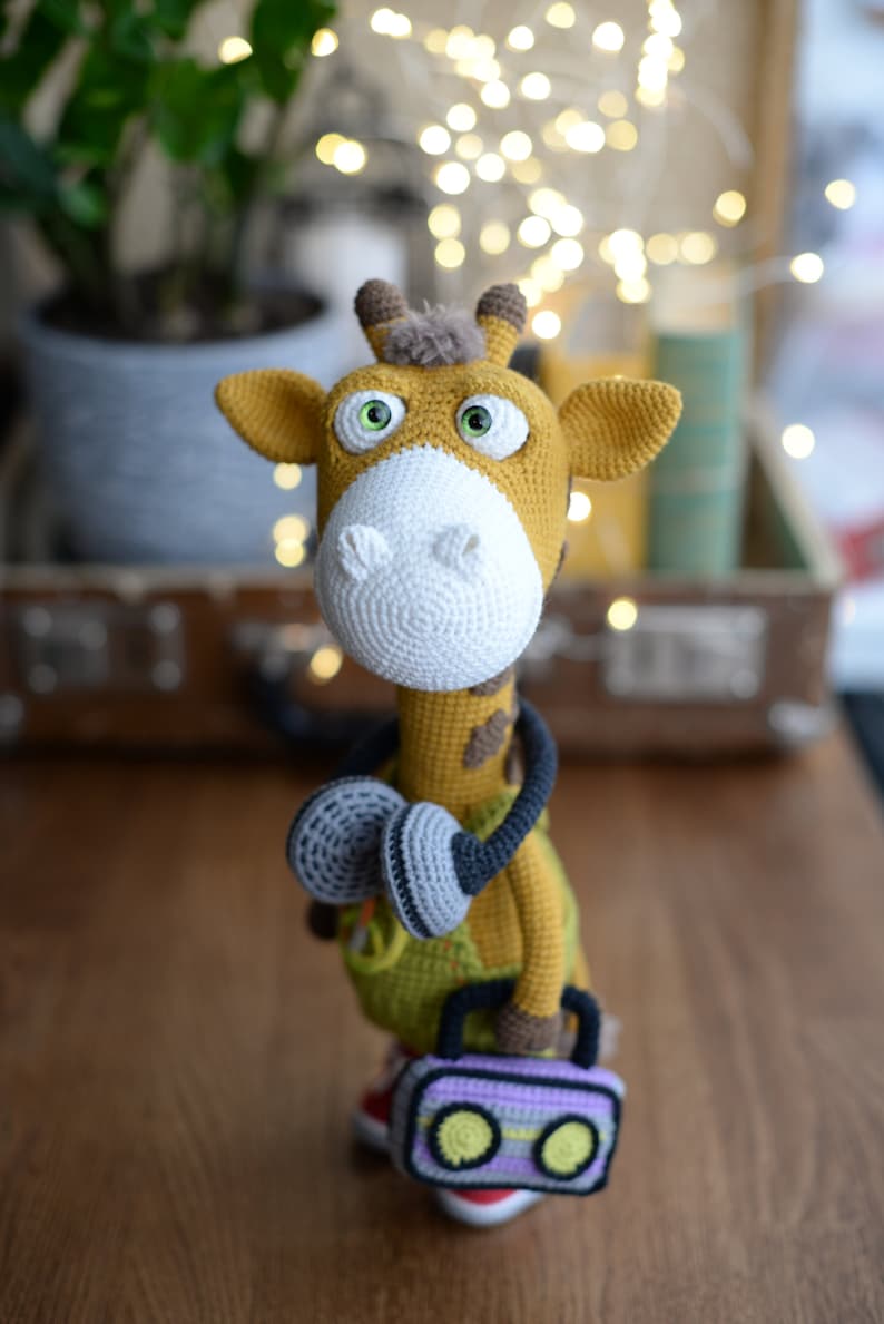 May include: A crocheted giraffe toy wearing headphones and holding a purple and yellow boombox. The giraffe has brown spots and green clothing.