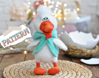 May include: A white crocheted goose with a blue and green hooded sweater, orange beak and feet, and blue eyes. The goose is sitting on a woven straw mat.