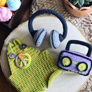 May include: Crocheted toy set featuring a green and yellow peace sign overalls, a purple and gray cassette tape, and a pair of gray and black headphones. The toys are arranged on a wooden surface with yarn balls and a potted succulent in the background.
