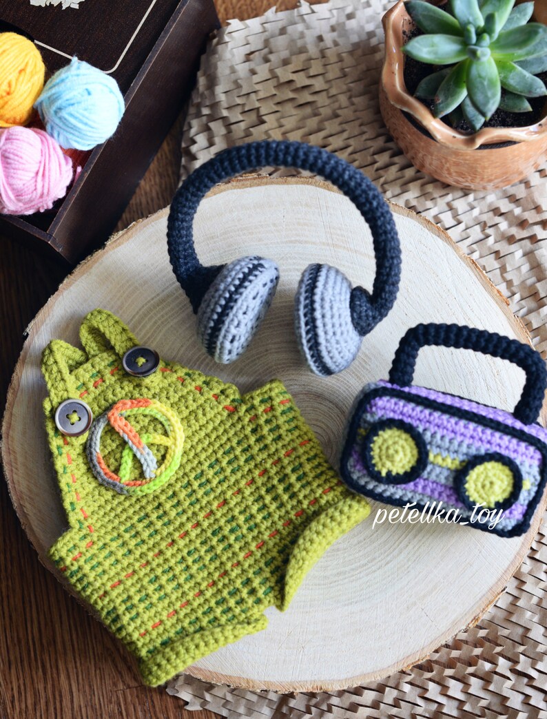 May include: Crocheted toy set featuring a green and yellow peace sign overalls, a purple and gray cassette tape, and a pair of gray and black headphones. The toys are arranged on a wooden surface with yarn balls and a potted succulent in the background.