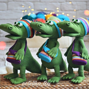 May include: Three green crocheted alligator toys wearing colorful hats and carrying small crocheted bags with rainbow stripes. The alligators are standing on a woven basket.