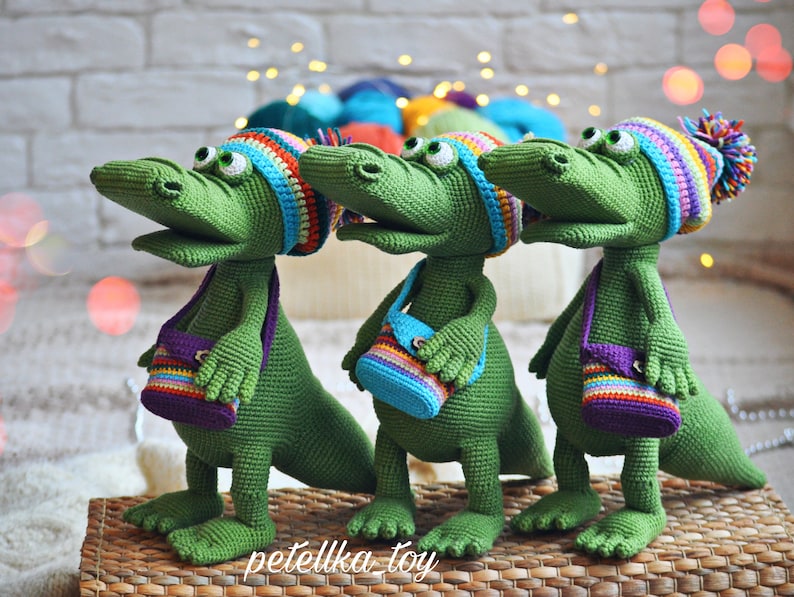 May include: Three green crocheted alligator toys wearing colorful hats and carrying small crocheted bags with rainbow stripes. The alligators are standing on a woven basket.