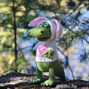 May include: A green crocheted dinosaur wearing a white bunny costume with pink ears and a pink heart on the front. The dinosaur is standing on a rock covered in moss.