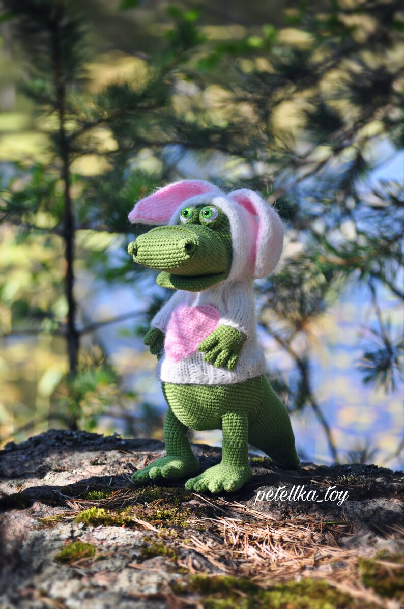 May include: A green crocheted dinosaur wearing a white bunny costume with pink ears and a pink heart on the front. The dinosaur is standing on a rock covered in moss.
