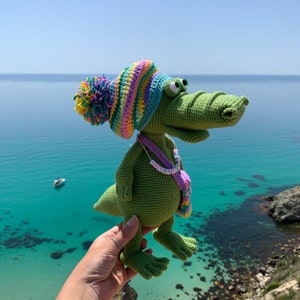 May include: A green crocheted alligator wearing a colorful striped hat with a pom-pom. The alligator is holding a small purple and yellow bag with a beaded necklace that says "GET LIT" in colorful letters.