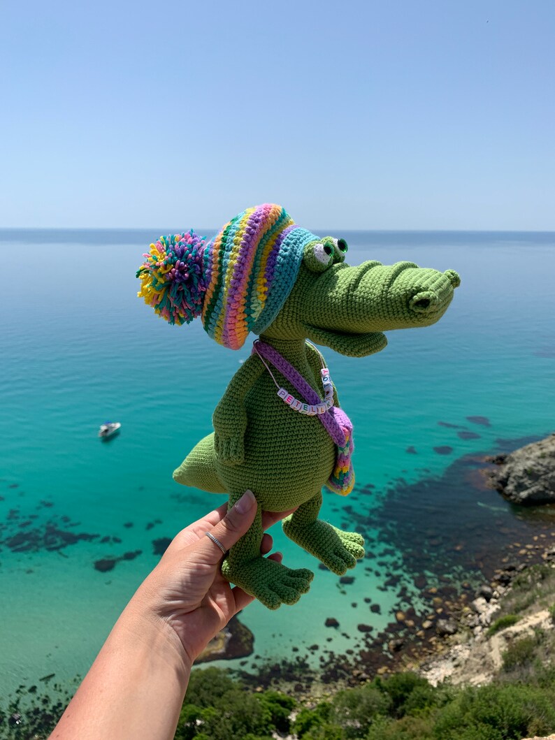 May include: A green crocheted alligator wearing a colorful striped hat with a pom-pom. The alligator is holding a small purple and yellow bag with a beaded necklace that says "GET LIT" in colorful letters.