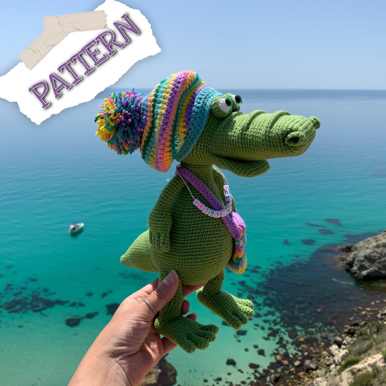 May include: A green crocheted alligator wearing a colorful striped hat and a purple beaded necklace with the word "Petite" spelled out in white beads. The alligator is holding a small purple crocheted bag.