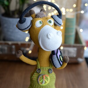 May include: A crocheted giraffe wearing headphones and overalls with a peace sign. The giraffe is standing on a wooden surface.