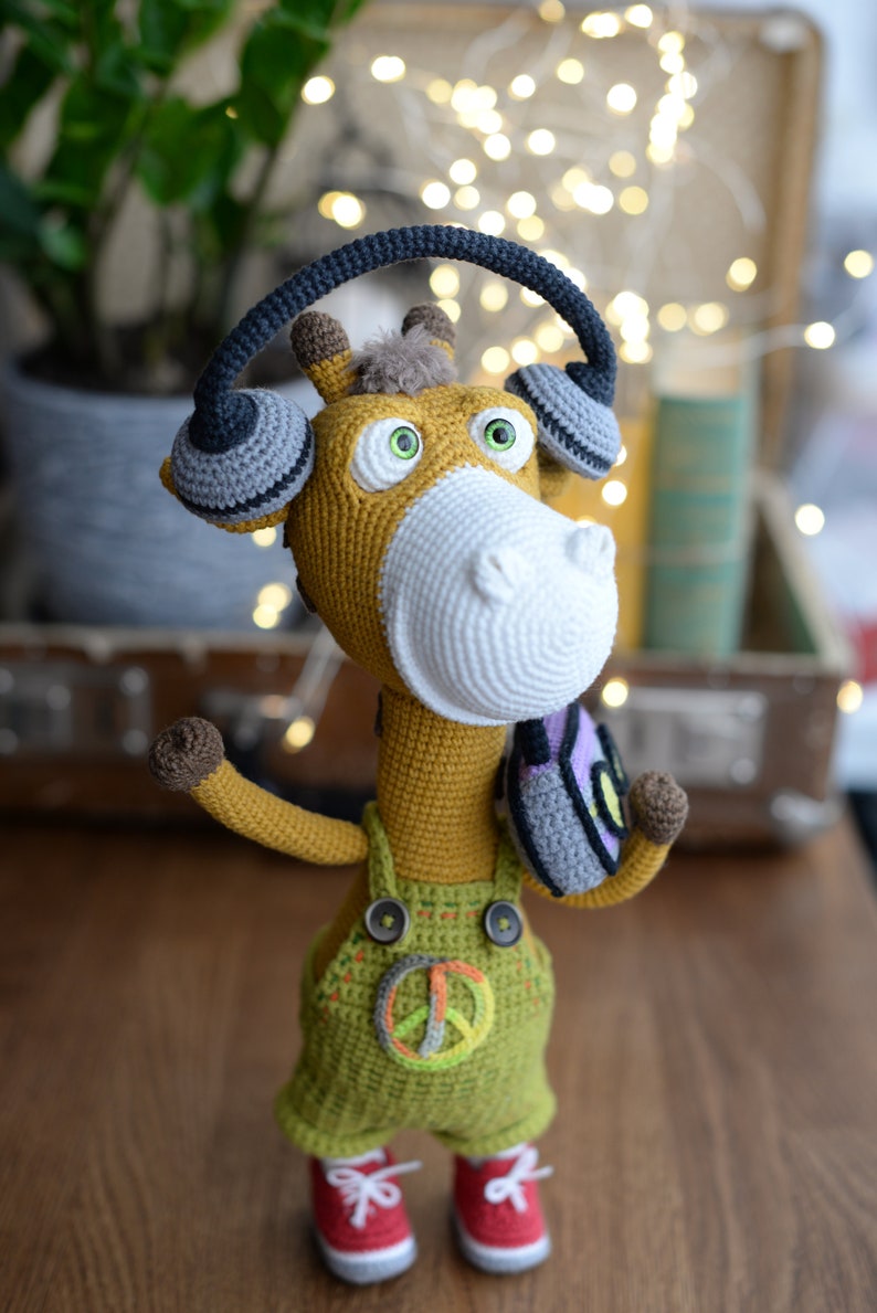 May include: A crocheted giraffe wearing headphones and overalls with a peace sign. The giraffe is standing on a wooden surface.