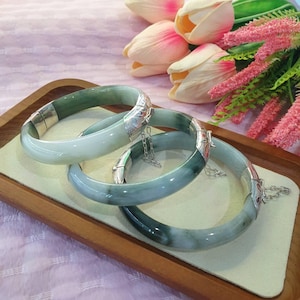May include: Three jade bangles with silver clasps rest on a wooden tray. The jade varies in shades of green and white. The clasps have intricate designs and are connected by small silver chains. The background includes pink and white flowers.
