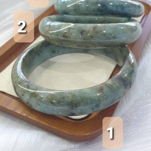 May include: Three jade bangles in varying shades of green and brown are displayed on a wooden tray. The bangles have a smooth, polished surface and are arranged in a stack, showcasing their natural color variations.
