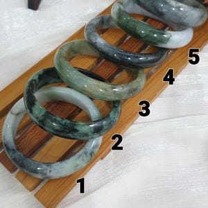 May include: Six smooth, polished jade bangles arranged in a row on a wooden rack, numbered one through six. Each bangle features unique natural variations in green, white, and dark gray tones, showcasing a translucent, rounded, and circular design.