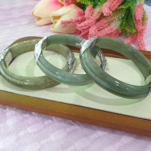 Può includere: Tre braccialetti in giada verde traslucida con dettagli argentati sono esposti su un vassoio di legno. Le chiusure argentate aggiungono un tocco di eleganza. I braccialetti sono un accessorio classico e senza tempo.