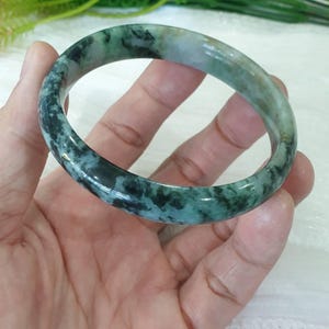 Puede incluir: Un brazalete de jade con un patrón moteado verde y negro. La superficie lisa y pulida refleja la luz, mostrando las variaciones naturales de color. El brazalete tiene forma circular, diseñado para ser usado en la muñeca.